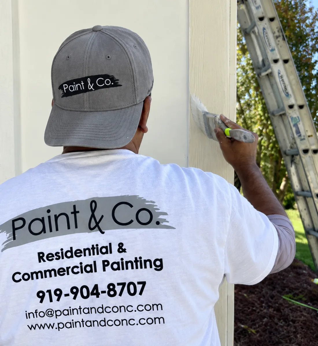 Painter in a Paint & Co. shirt brushing paint onto exterior trim beside a ladder