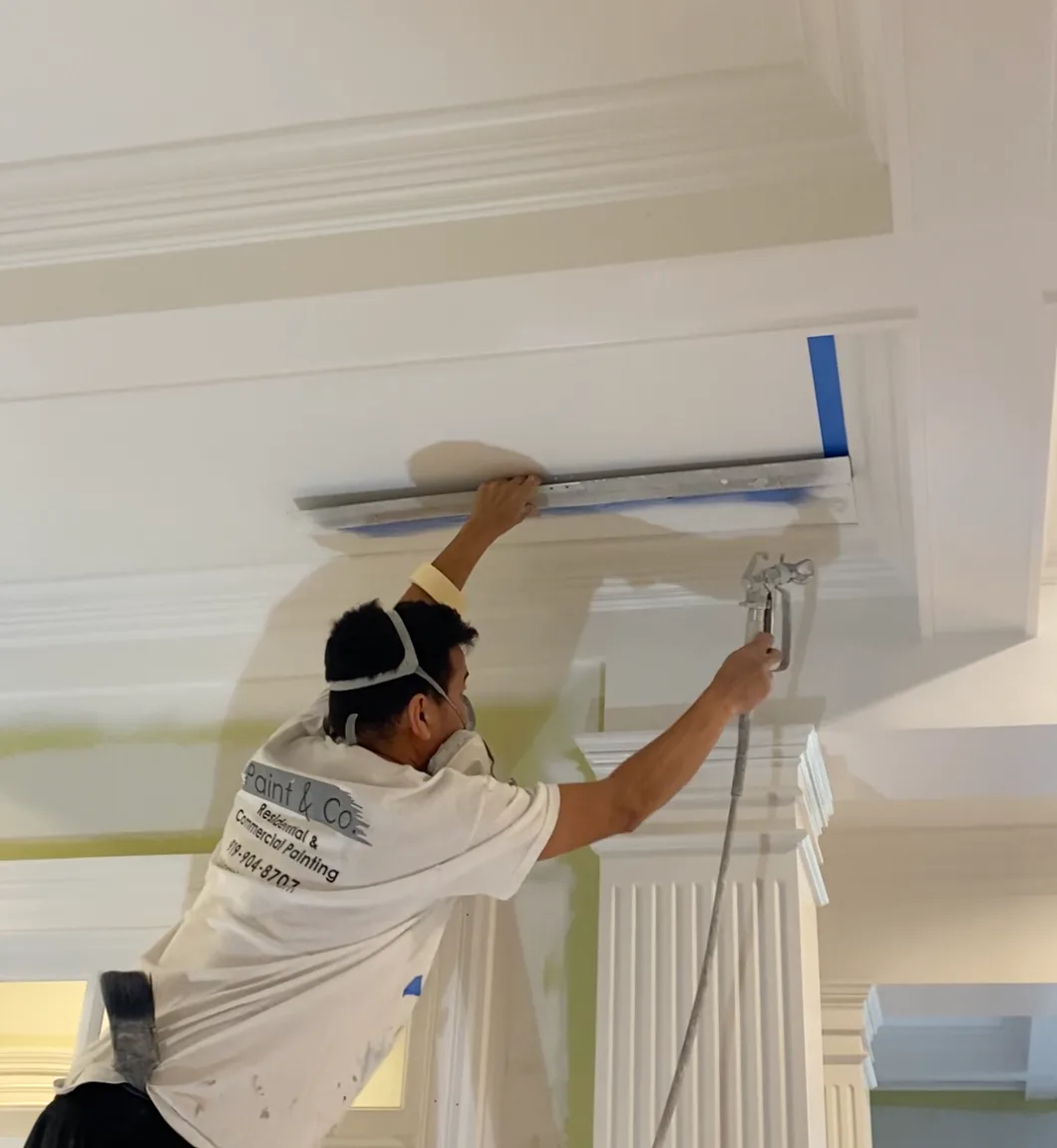 Painter in a respirator spraying a coffered ceiling and column inside a home