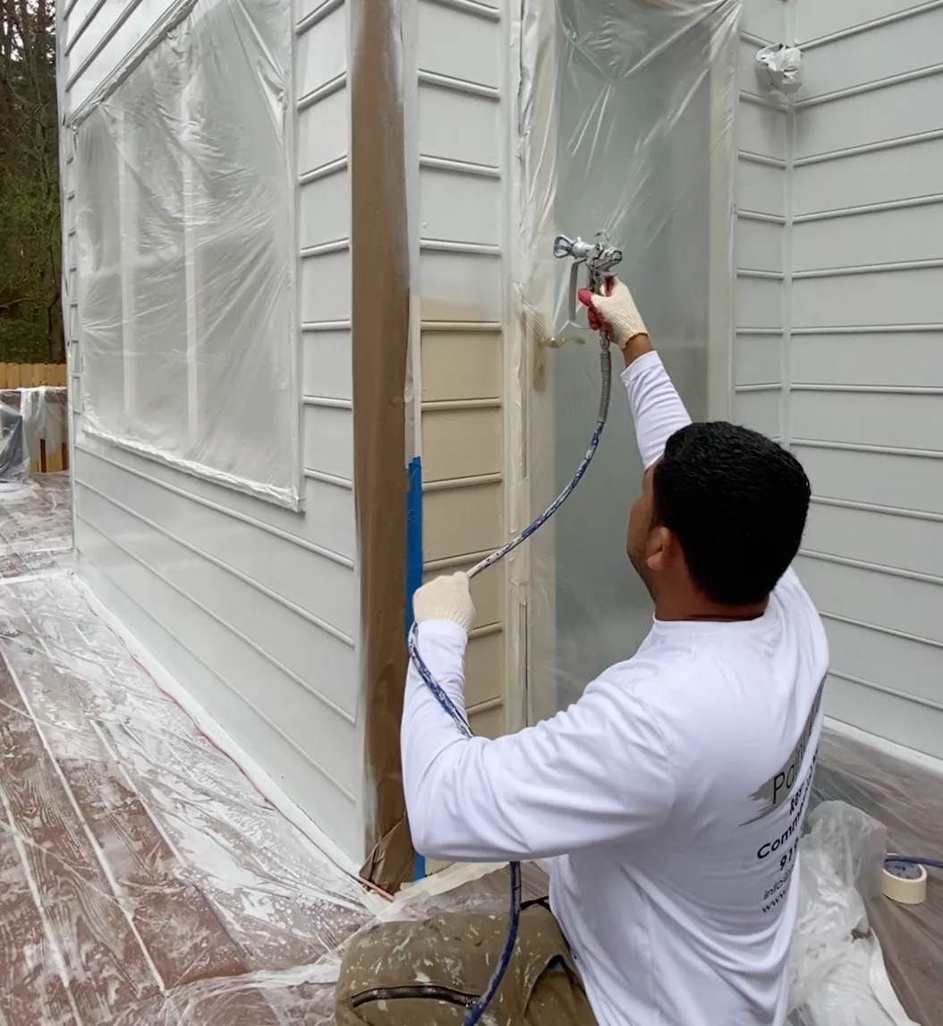 Painter spraying white exterior siding with the windows masked in plastic
